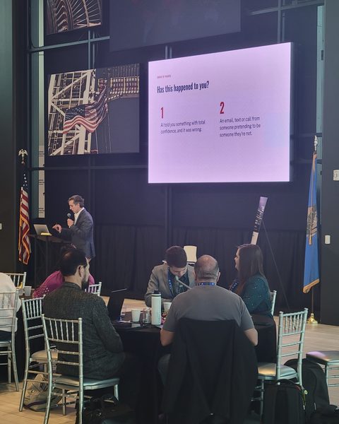 Matt Bertram at the lectern in front of the symposium audience, beside a slide headed Has this happened to you, asking whether AI has told the room something wrong with total confidence, or someone has pretended to be someone they are not